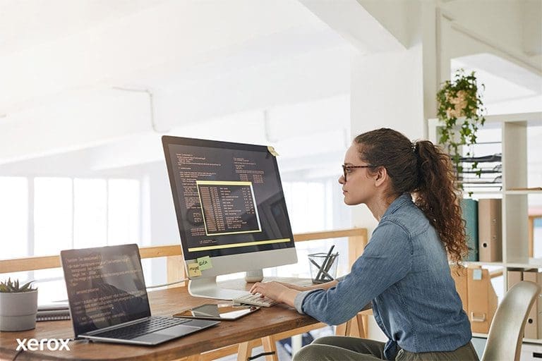 Una mujer sentada en su escritorio, con una computadora y una laptop, demostrando productividad en un espacio de trabajo moderno.