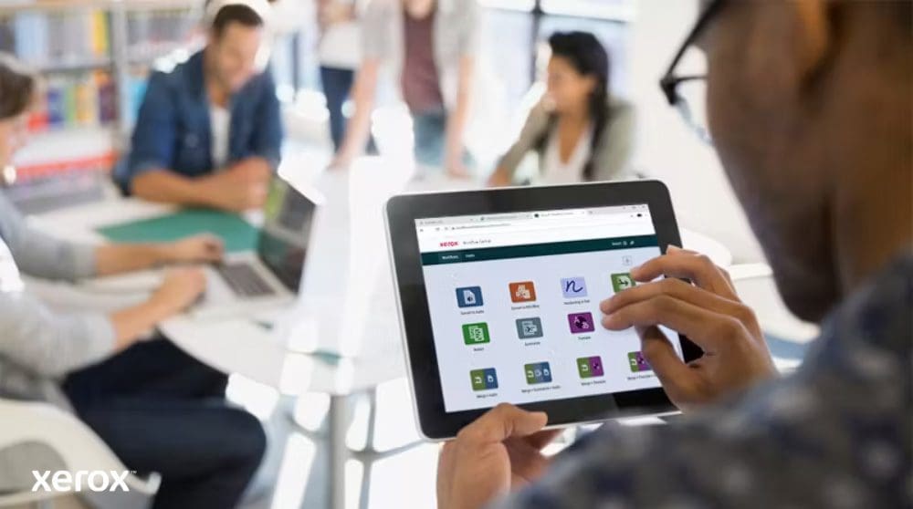Una persona sostiene una tableta que muestra la interfaz de Xerox Workflow Central con varios íconos de aplicaciones, mientras otras personas trabajan en el fondo borroso, resaltando el acceso móvil a las soluciones de flujo de trabajo.