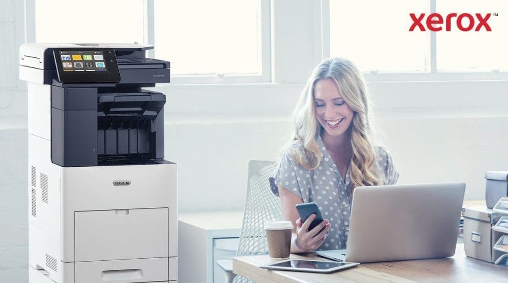 Una persona mira su smartphone con satisfacción, sentada en un escritorio con una laptop. Junto al escritorio, en una oficina bien iluminada, se ve una impresora multifunción Xerox VersaLink B605.