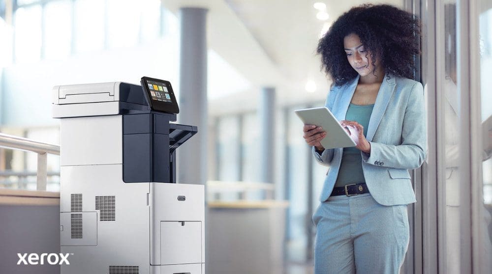Una persona se encuentra junto a una impresora multifunción Xerox VersaLink B605, mirando e interactuando con una tableta, lo que ilustra cómo ConnectKey permite la interacción móvil con la impresora.