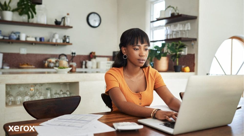 Una persona está sentada en una mesa de cocina, concentrada en trabajar en una computadora portátil, con papeles y una calculadora cerca, lo que ilustra la flexibilidad de trabajar desde casa con una gestión eficiente de documentos.