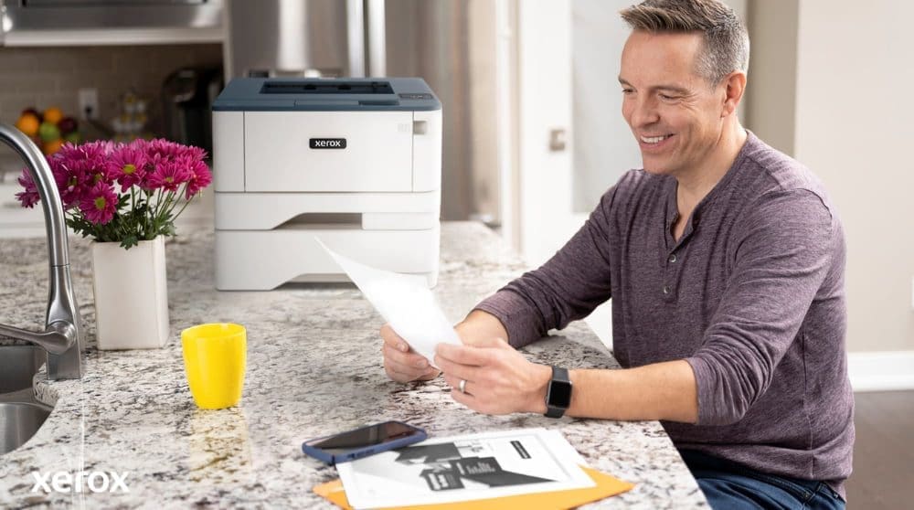 Una persona sonríe mientras revisa un documento en el mostrador de la cocina, con una impresora Xerox compacta y una taza de color amarillo brillante cerca, lo que sugiere una impresión y un manejo de documentos sin inconvenientes en un hogar o una pequeña oficina.