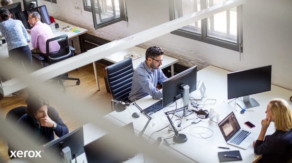 Vista aérea de una oficina diáfana con varias personas trabajando en sus escritorios y usando computadoras. La escena transmite un ambiente de trabajo productivo y colaborativo.