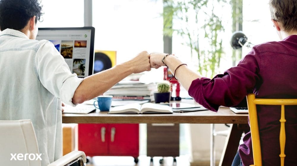 Dos personas sentadas en un escritorio, de espaldas a la cámara, chocan los puños, símbolo del trabajo en equipo y la colaboración exitosa. Una mira la pantalla de una computadora.