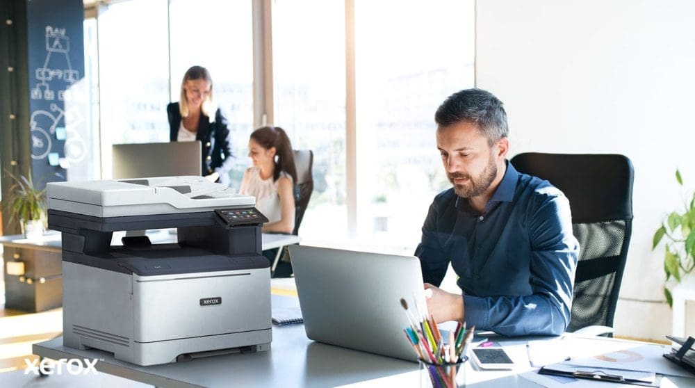 Una persona está sentada en un escritorio, mirando una computadora portátil con una impresora multifunción Xerox C325 al lado. Al fondo, otras dos personas también trabajan en sus escritorios en un ambiente de oficina bien iluminado.